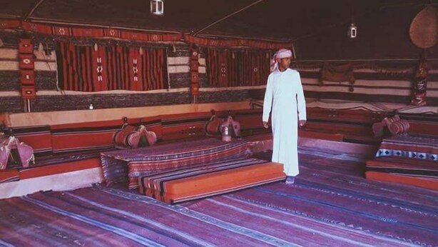 A traditional room with a patterned floor, wooden benches, and a person standing. - Bedouin Experience Camp (Wadi Rum)