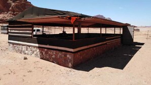 A rustic outdoor seating area with a stone base and a metal roof, surrounded by a desert landscape. - Bedouin Experience Camp (Wadi Rum)