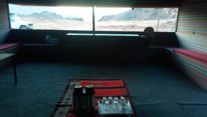 A room with a large window, a red rug, a table with a kettle and cups, and a bench. - Bedouin Experience Camp (Wadi Rum)