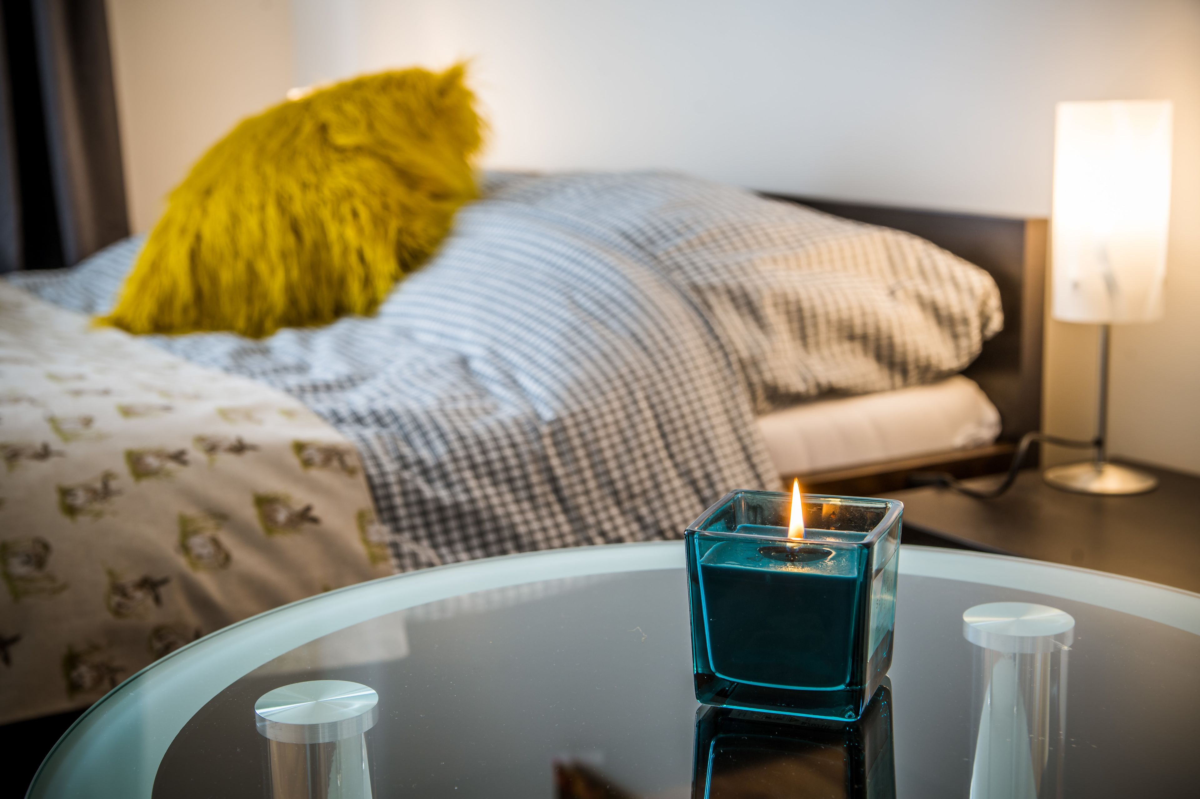 A neatly made bed with a patterned bedspread and a single yellow pillow. A teal candle sits on a glass table in front of the bed.