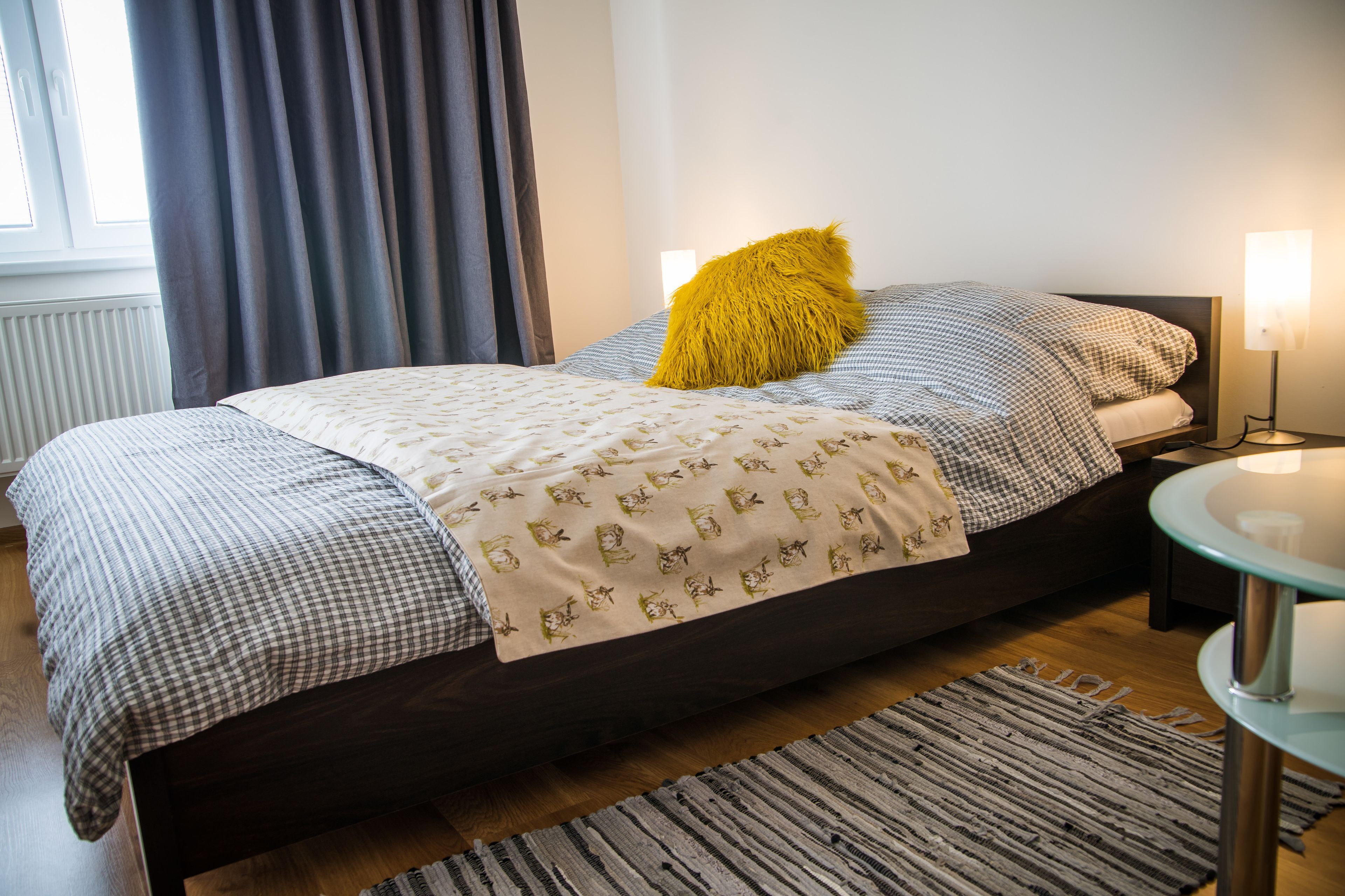 Una cama bien hecha con una colcha a cuadros, un cojín amarillo y una manta estampada. Se ve una mesita de noche con una lámpara, una alfombra en el suelo y una ventana con cortinas.