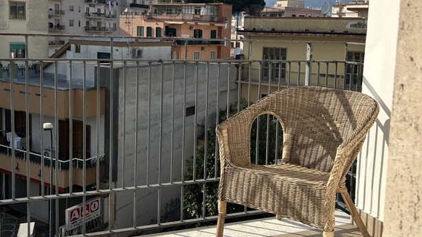 Un balcón con una silla de mimbre, baranda metálica y vistas a edificios y montañas al fondo.