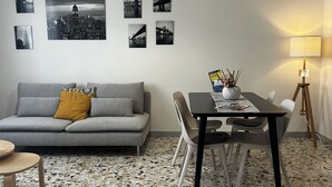 Una sala moderna con un sofá gris, una mesa de comedor con sillas, una lámpara de pie y una obra de arte con vistas a la ciudad en la pared.