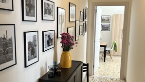 Un pasillo con varias fotografías enmarcadas en la pared, un jarrón amarillo con flores sobre un mueble y una silla en primer plano.