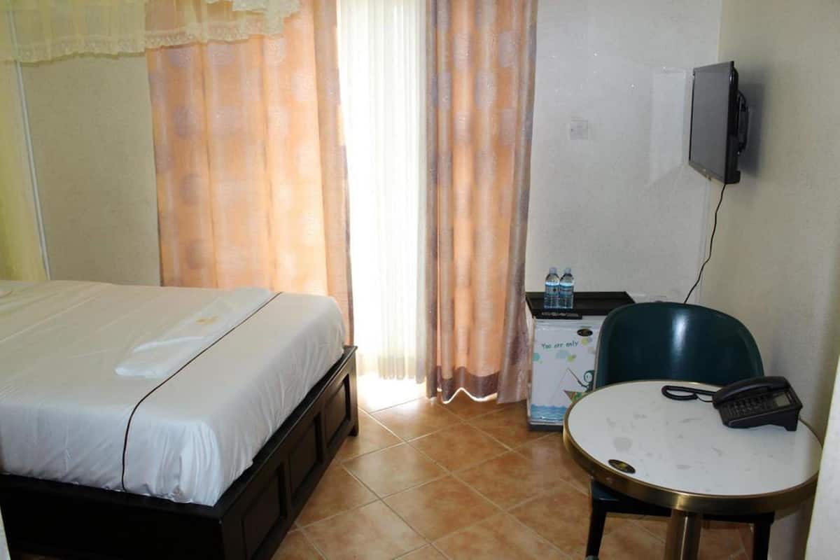 Quarto de hotel com uma cama, uma escrivaninha com telefone, uma televisão na parede e um pequeno armário com dispensador de água.