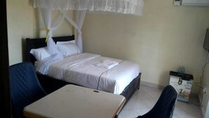 A single bed with a canopy, a small table, and a chair in a room with a tiled floor and a wall-mounted air conditioner. - Fancy Hotel (Kampala)