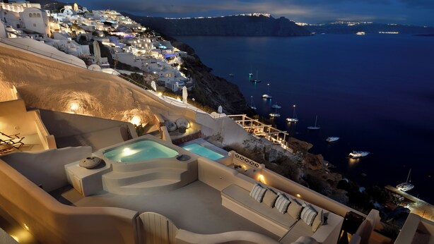 A luxurious outdoor area with a hot tub, seating, and a view of a coastal town at night. - Archon Villa by K&K (Oia)