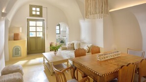 A dining area with a wooden table and chairs, a hanging light fixture, and a view of a kitchen and living area through an arched doorway. - Archon Villa by K&K (Oia)