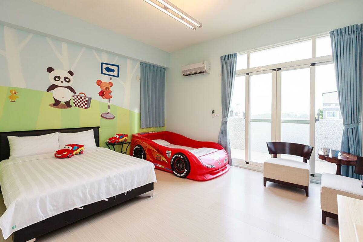 A bedroom with a bed, a red toy car, a panda and teddy bear wall decoration, a checkered bucket, and a large window with curtains.