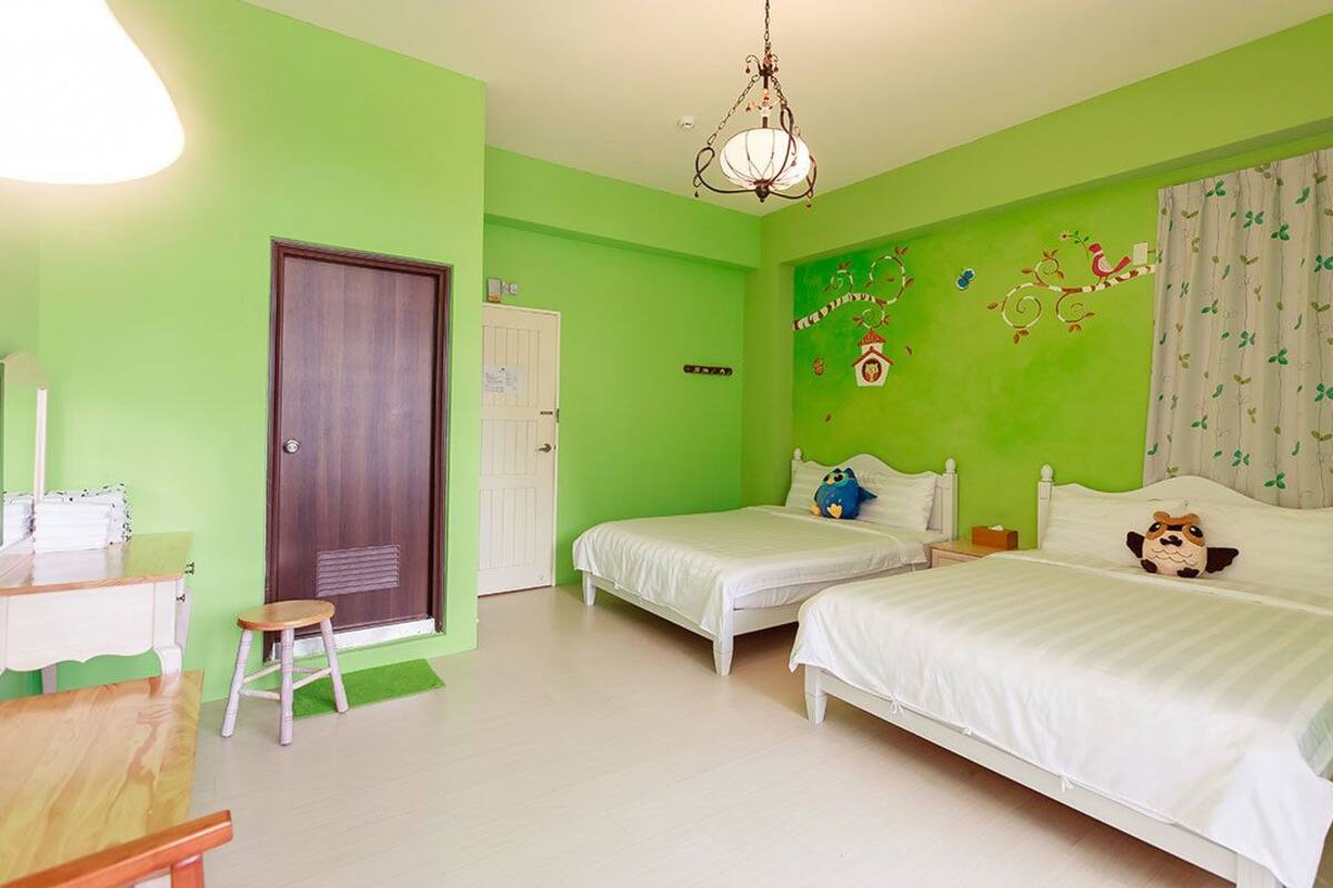 A room with two beds, a desk, and a chair. There is a door and a window with curtains.