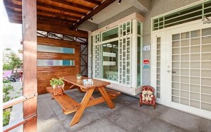 A covered outdoor area with a wooden table and benches, a potted plant, and a decorative chair.