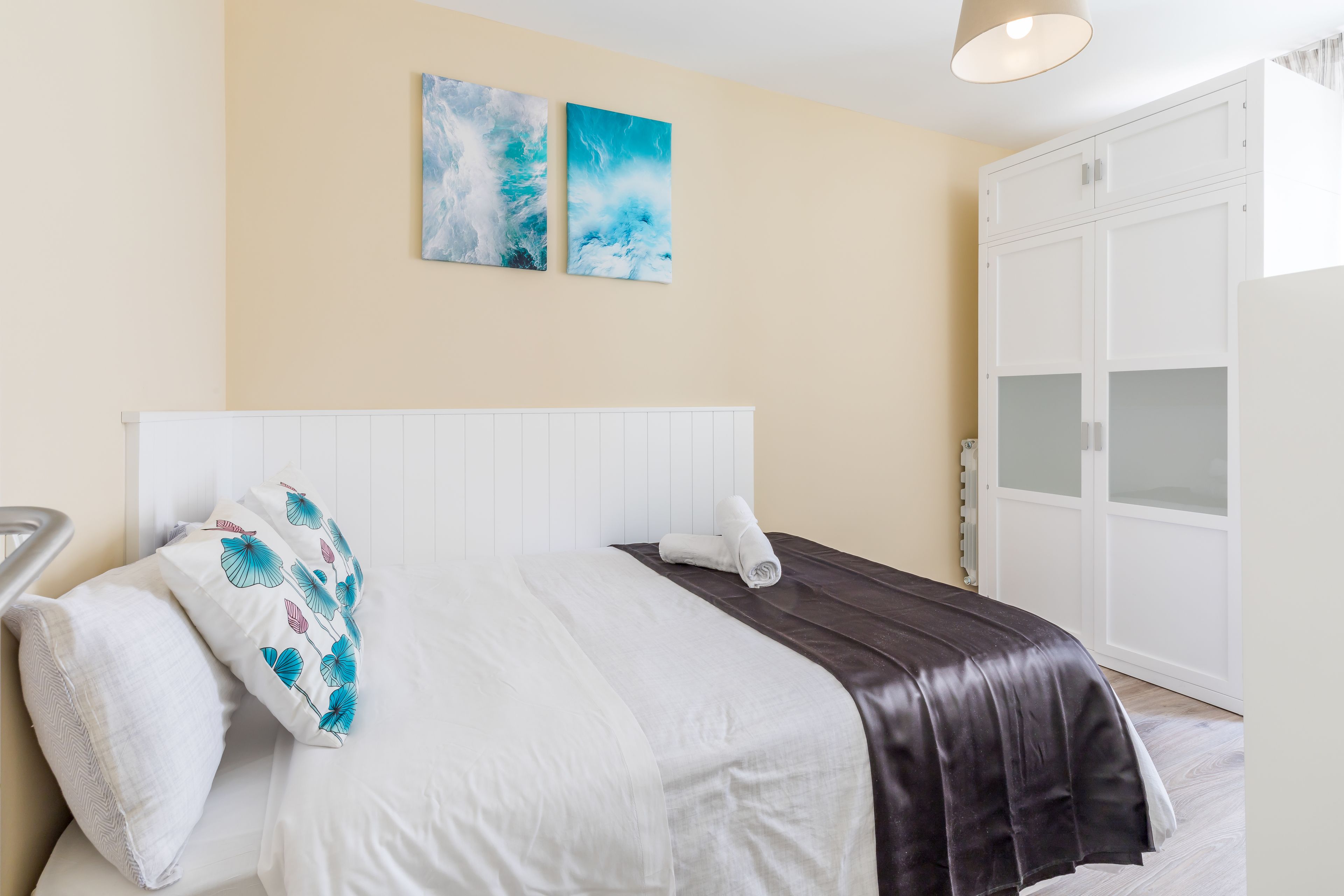 A neatly made bed with white and dark brown linens, a white headboard, and two wall-mounted artworks depicting abstract blue and white scenes.