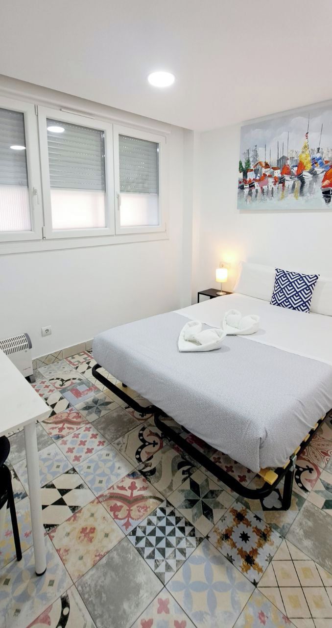 A hotel room with a bed, a bedside table, a chair, a window with blinds, a painting on the wall, and a patterned floor.