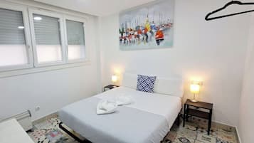 A bedroom with a bed, two bedside tables, a window with blinds, a painting on the wall, and a tiled floor.