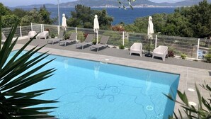 A pool area with lounge chairs and umbrellas, surrounded by a white fence, overlooking a scenic view of the sea and mountains.