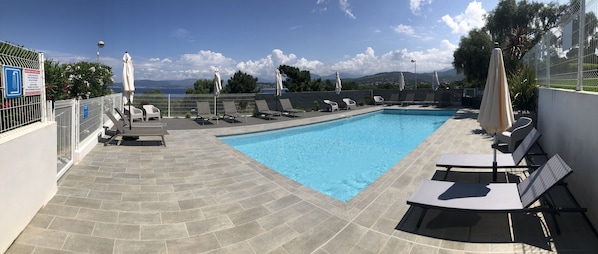 En pool med solstolar och parasoller, omgiven av ett staket och med utsikt över havet.