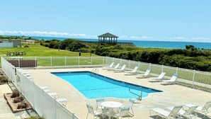 A rectangular swimming pool with lounge chairs and tables, surrounded by a white fence, with a gazebo and ocean view in the background. - Beachfront Community • Private Beach Access • Pool • WiFi • Kitchen (Atlantic Beach)