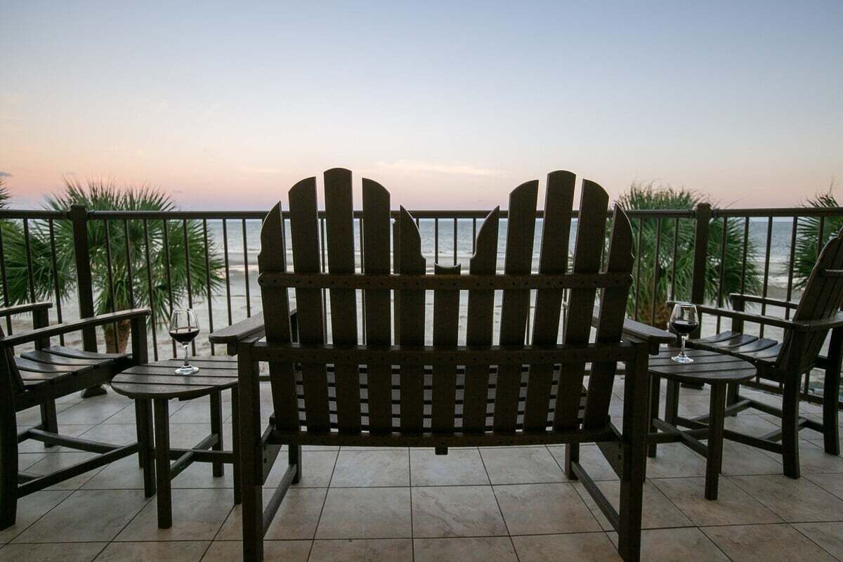 A wooden Adirondack chair and table set on a balcony overlooking the ocean.