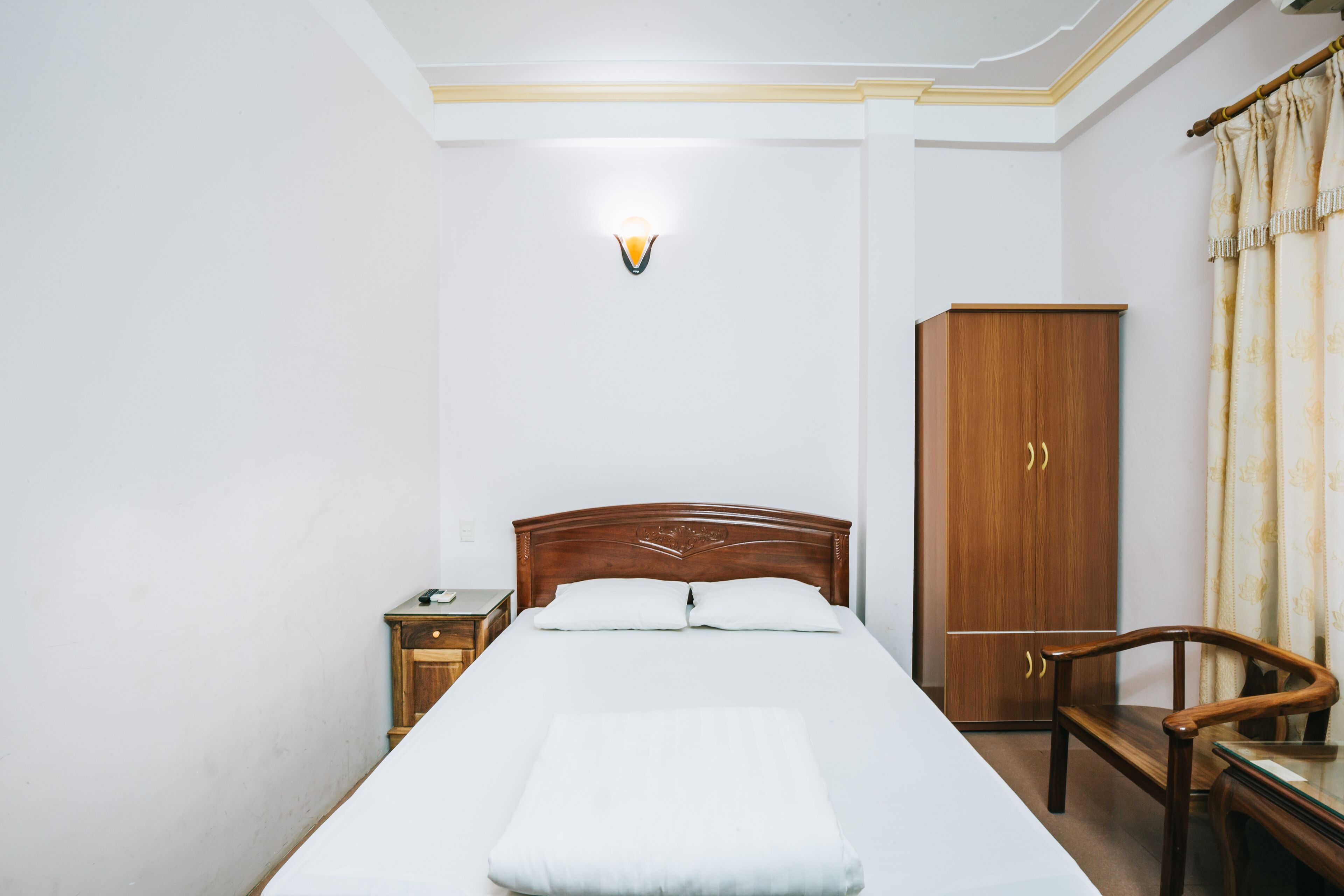 A neatly made bed with a white comforter and pillows, a wooden nightstand with a lamp, a wooden wardrobe, and a wooden chair.