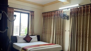 Ein Hotelzimmer mit einem Bett, Vorhängen, einem Ventilator und Blick auf Gebäude vor dem Fenster.