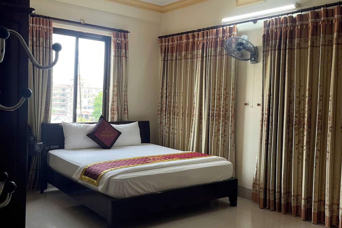A hotel room with a bed, curtains, a fan, and a view of buildings outside the window.