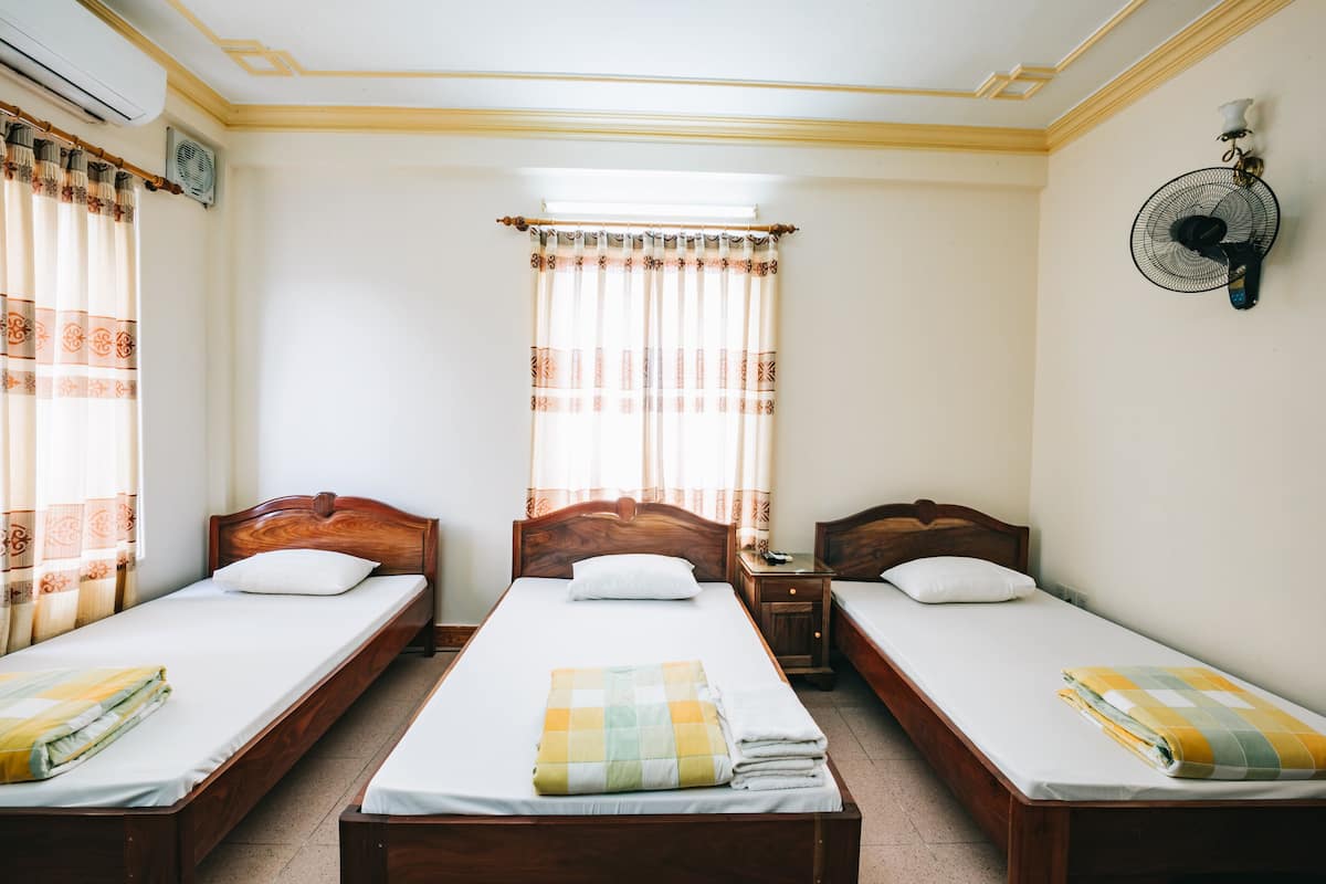 A room with three wooden beds, each with a white mattress and a folded blanket. There is a window with curtains, a wall-mounted fan, and a small table with a lamp.