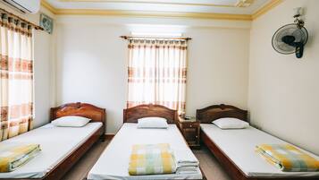 A room with three wooden beds, each with a white mattress and a folded blanket. There is a window with curtains, a wall-mounted fan, and a small table with a lamp.