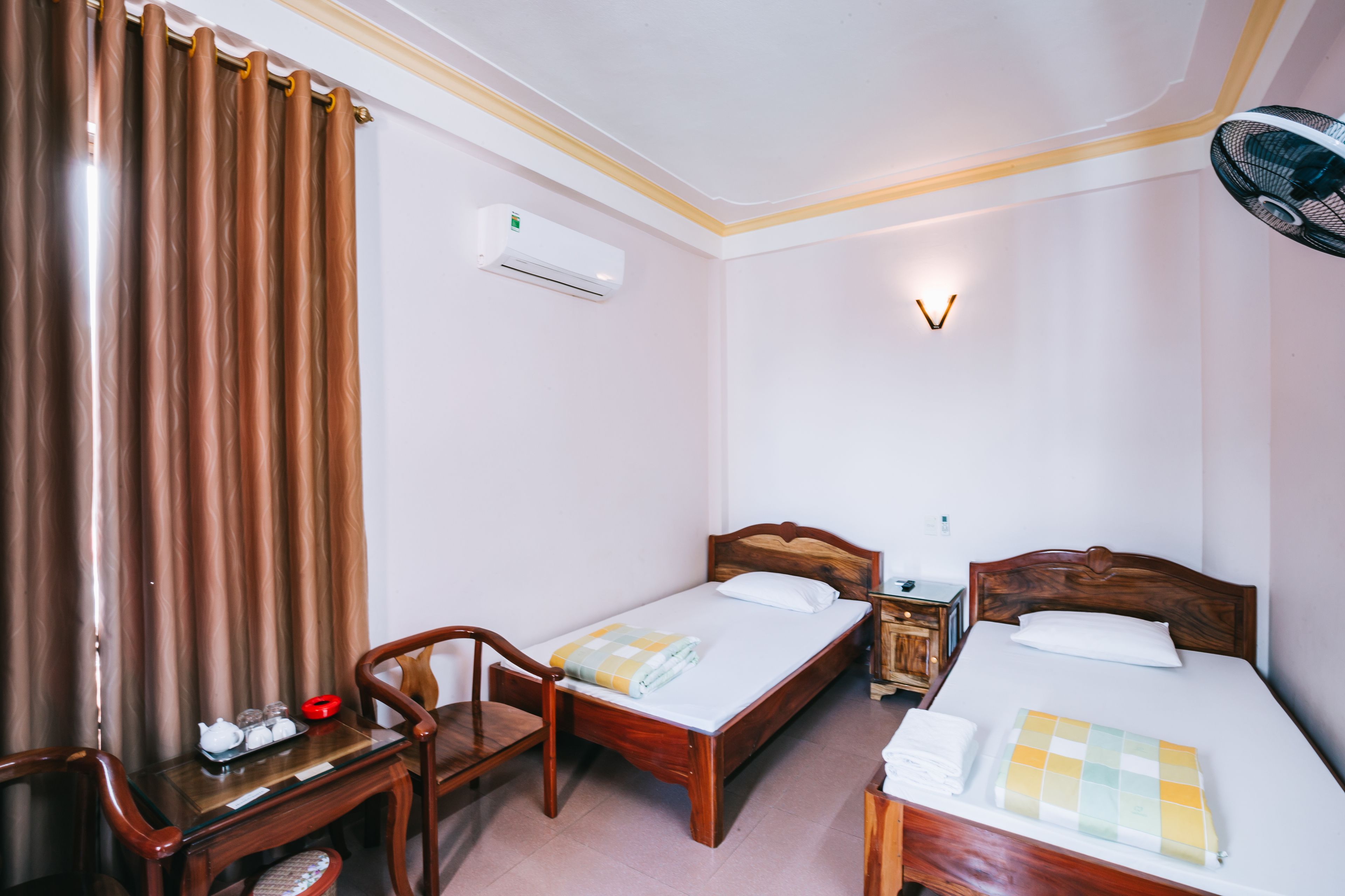 Une chambre d’hôtel avec deux lits simples, un bureau en bois avec une chaise, une table de chevet, un climatiseur et un ventilateur fixé au mur.