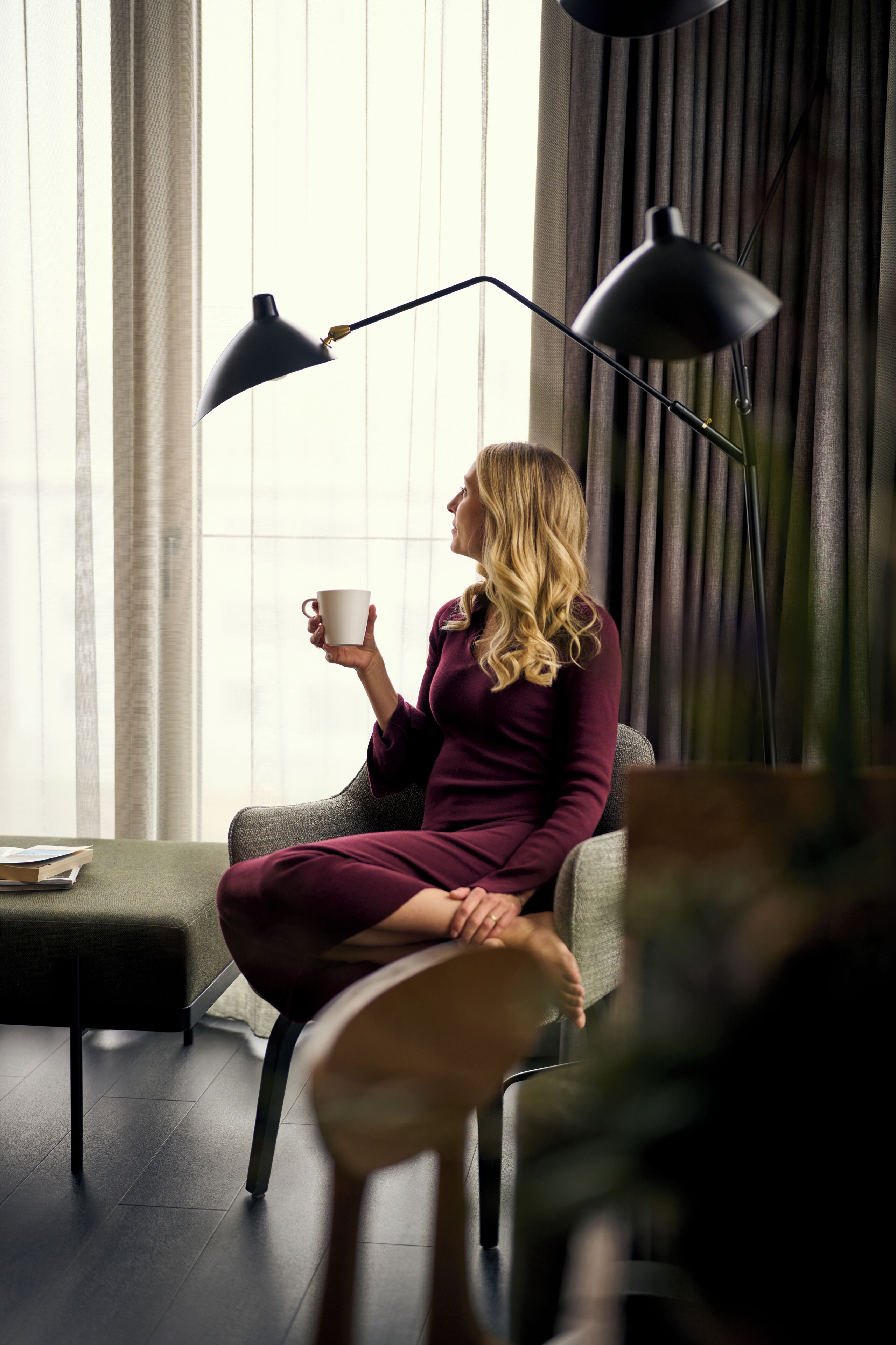 A person sitting on a grey sofa in a room with a large window, a black floor lamp, and a white mug.