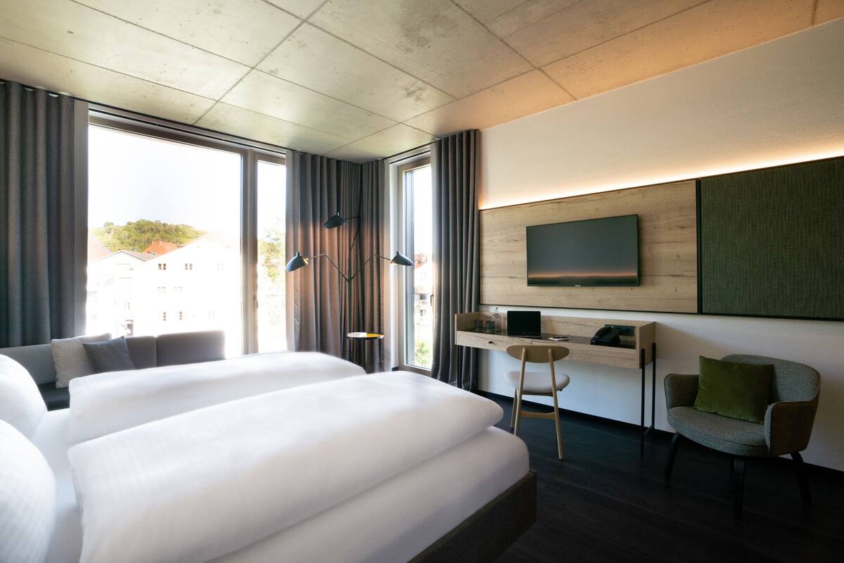Ein modernes Hotelzimmer mit zwei großen Betten, einem Schreibtisch mit Computer, einem Sessel und einem an der Wand befestigten Fernseher.