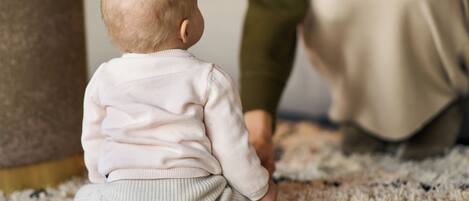 A baby sitting on a carpet with a woman standing behind.