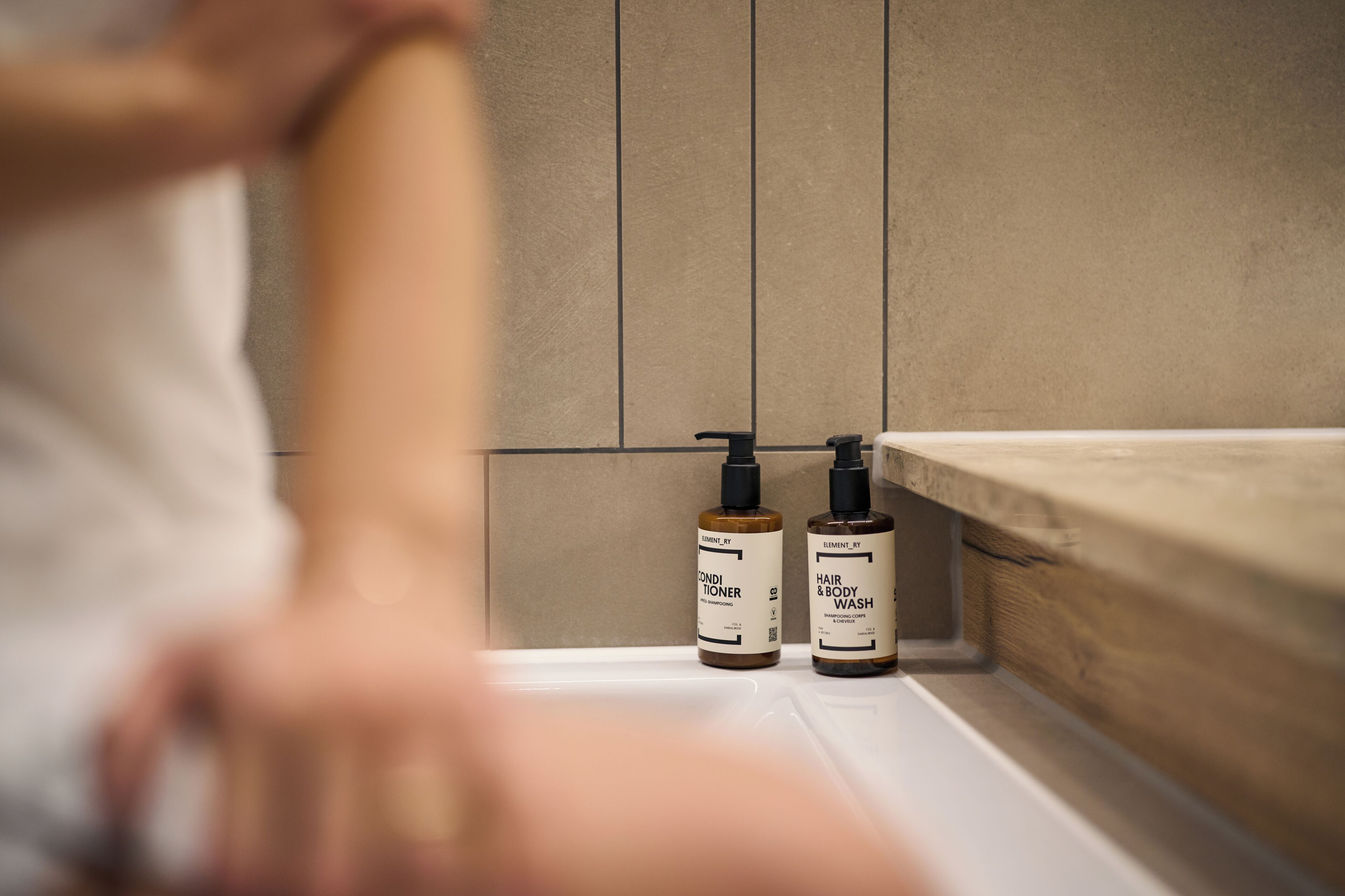 A bathroom with a wooden vanity, two bottles of hair and body wash, and a tiled wall.