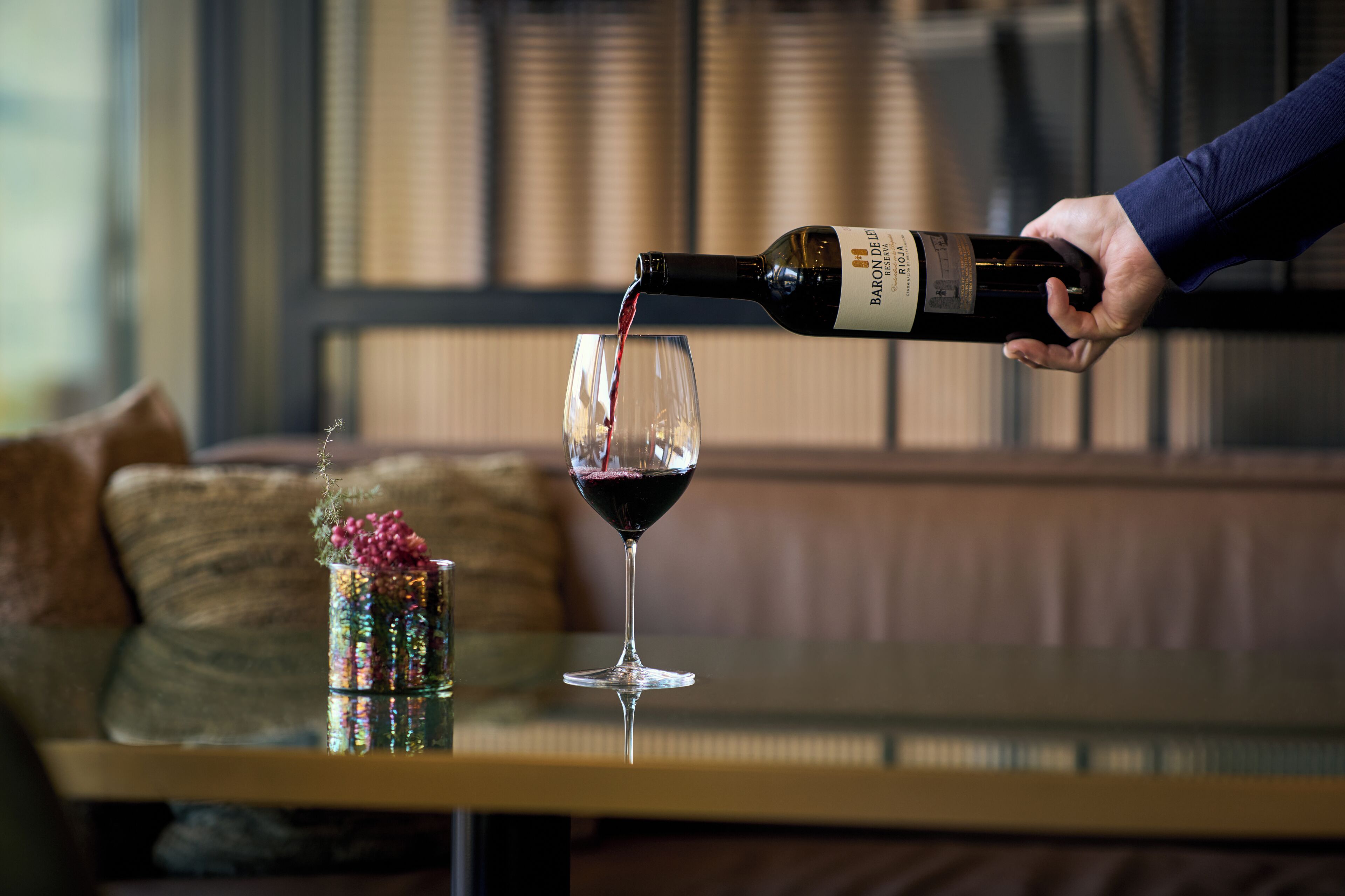 Una mano sirve vino en una copa sobre una mesa, con un sofá y cojines al fondo.