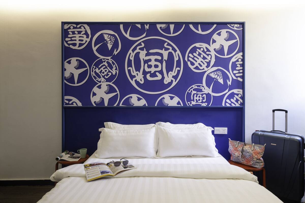 A hotel room with a bed, pillows, a magazine, a cup, a suitcase, and a wall with a blue and white pattern.