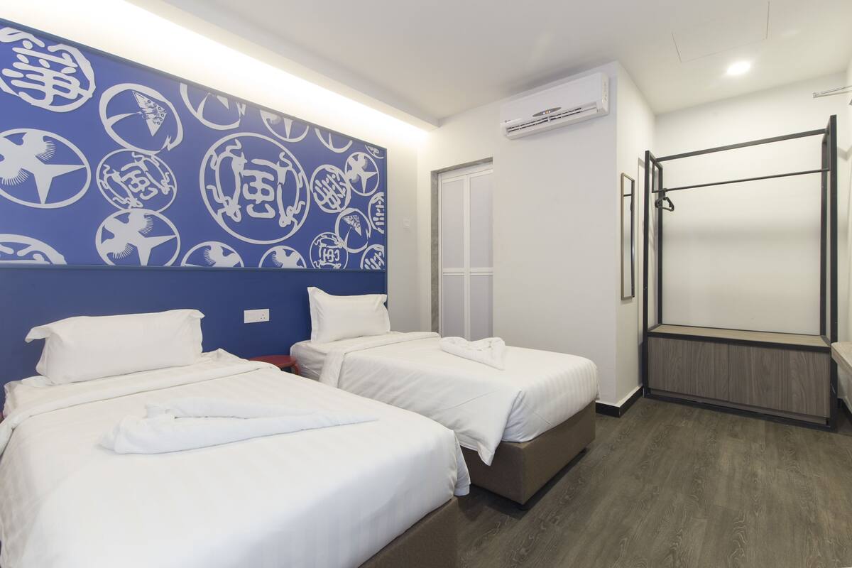 A hotel room with two beds, a blue wall with white patterns, a wooden floor, and a window.