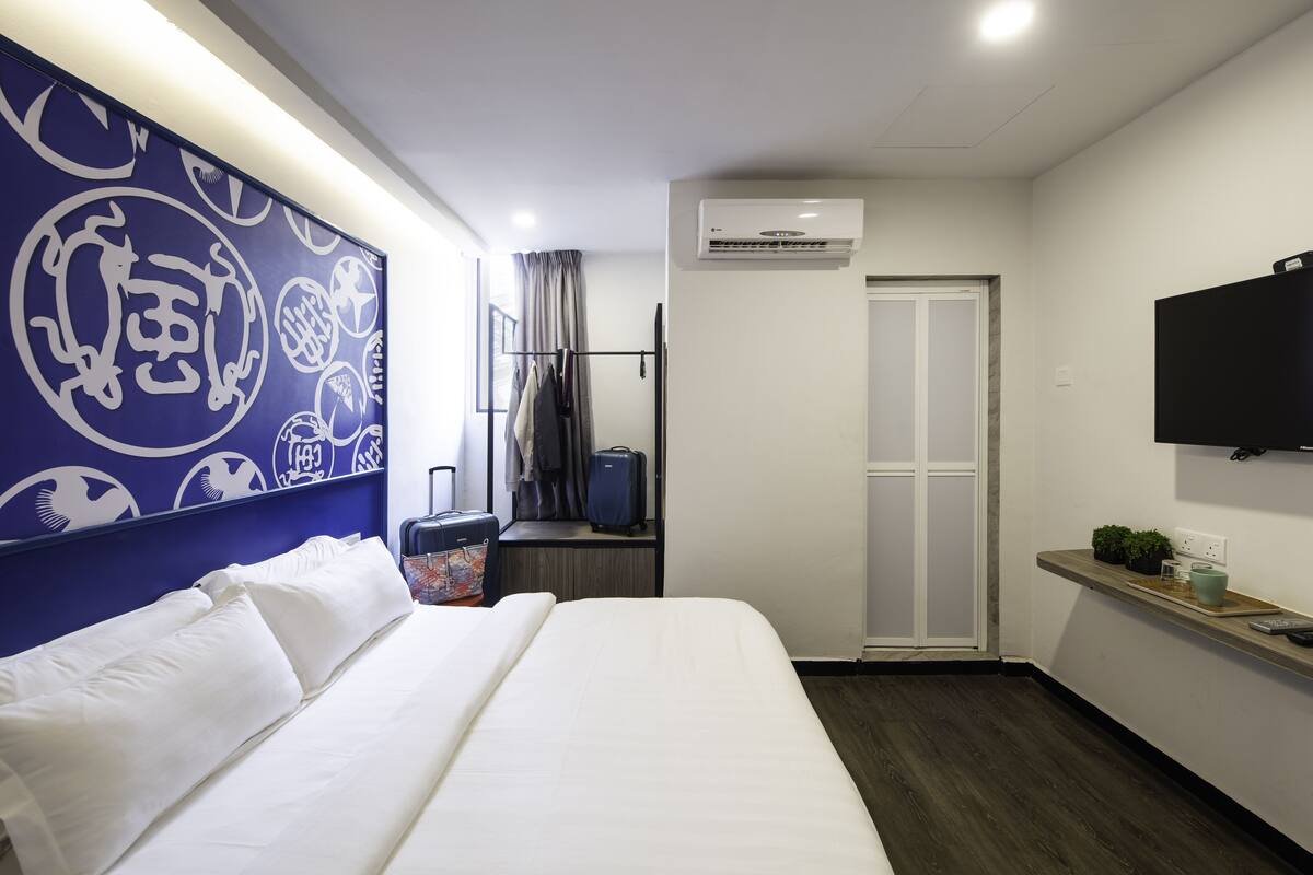 A hotel room with a bed, a wall-mounted TV, an air conditioning unit, a small table with a plant, and a window with curtains.