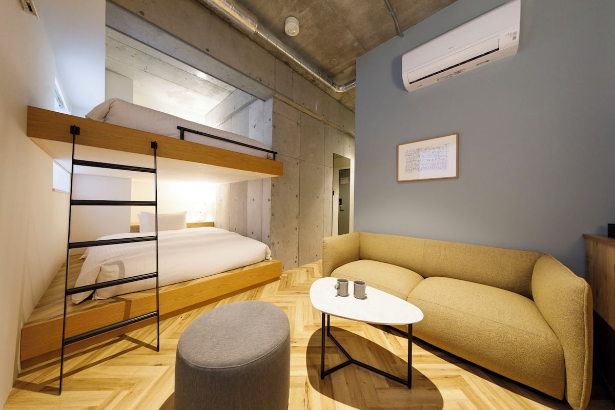 A compact hotel room with a bunk bed, a sofa, a small table, and an air conditioning unit.