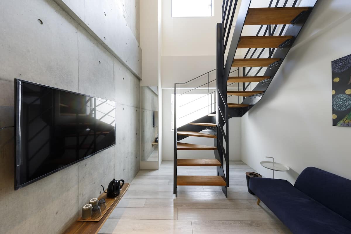 A modern interior with a flat-screen TV mounted on a concrete wall, a wooden staircase, a blue sofa, and a small table with cups and a kettle.