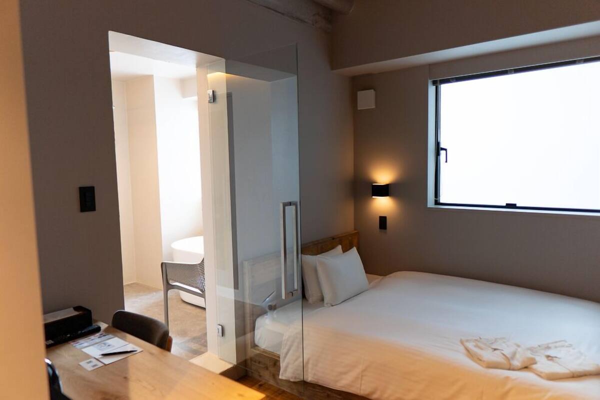 A hotel room with a bed, a desk with a chair, a window, and a door leading to another room.