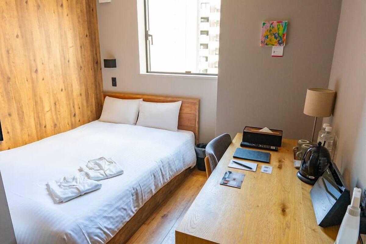 A hotel room with a bed, a desk with a lamp, a trash bin, and a window with a view of buildings.