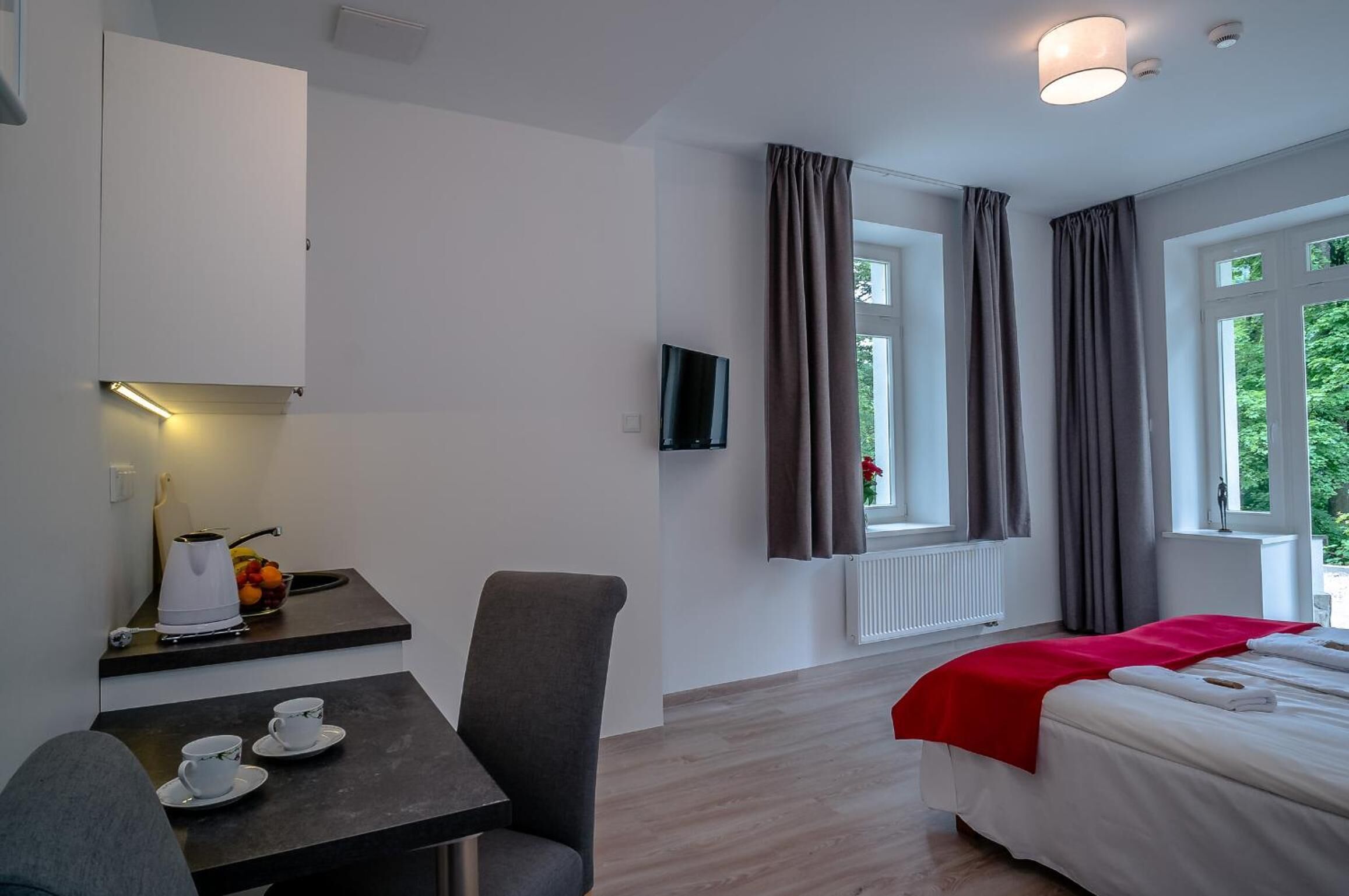A hotel room with a bed, a desk with a kettle and cups, a TV, and large windows with curtains.