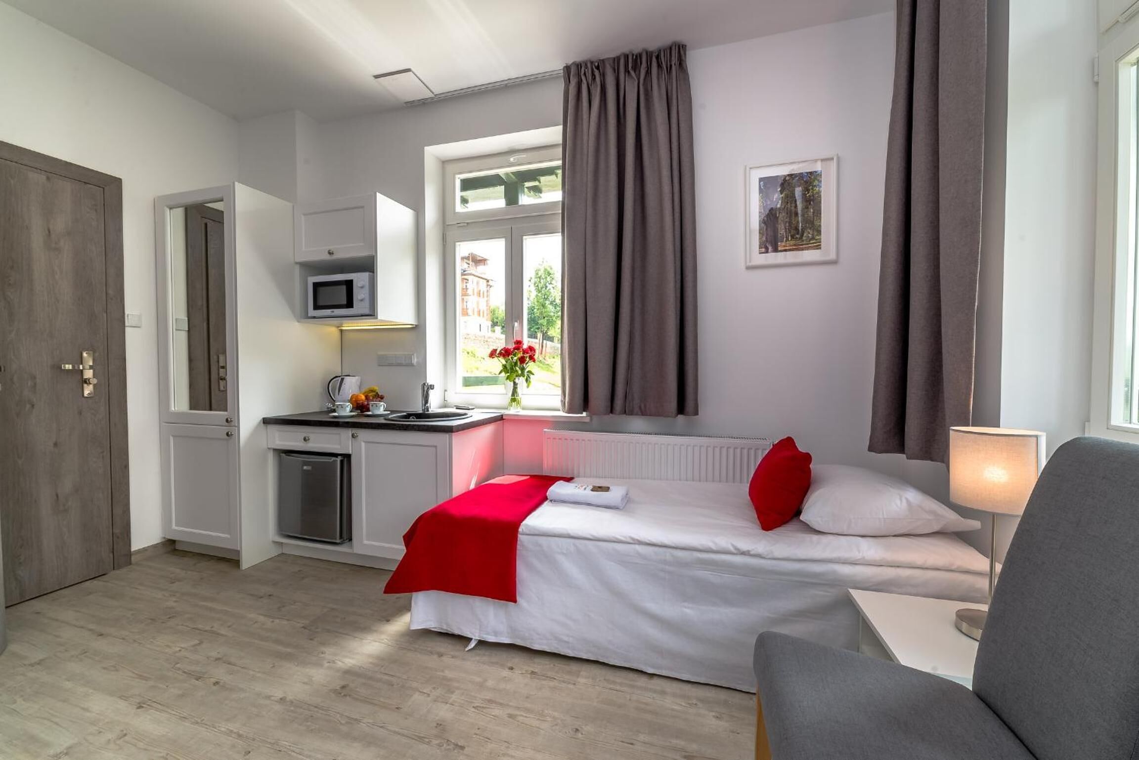 Ett kompakt hotellrum med ett litet pentry, en säng med rött och vitt sänglinne, ett fönster med gardiner, ett litet bord med en lampa och en tavla på väggen.
