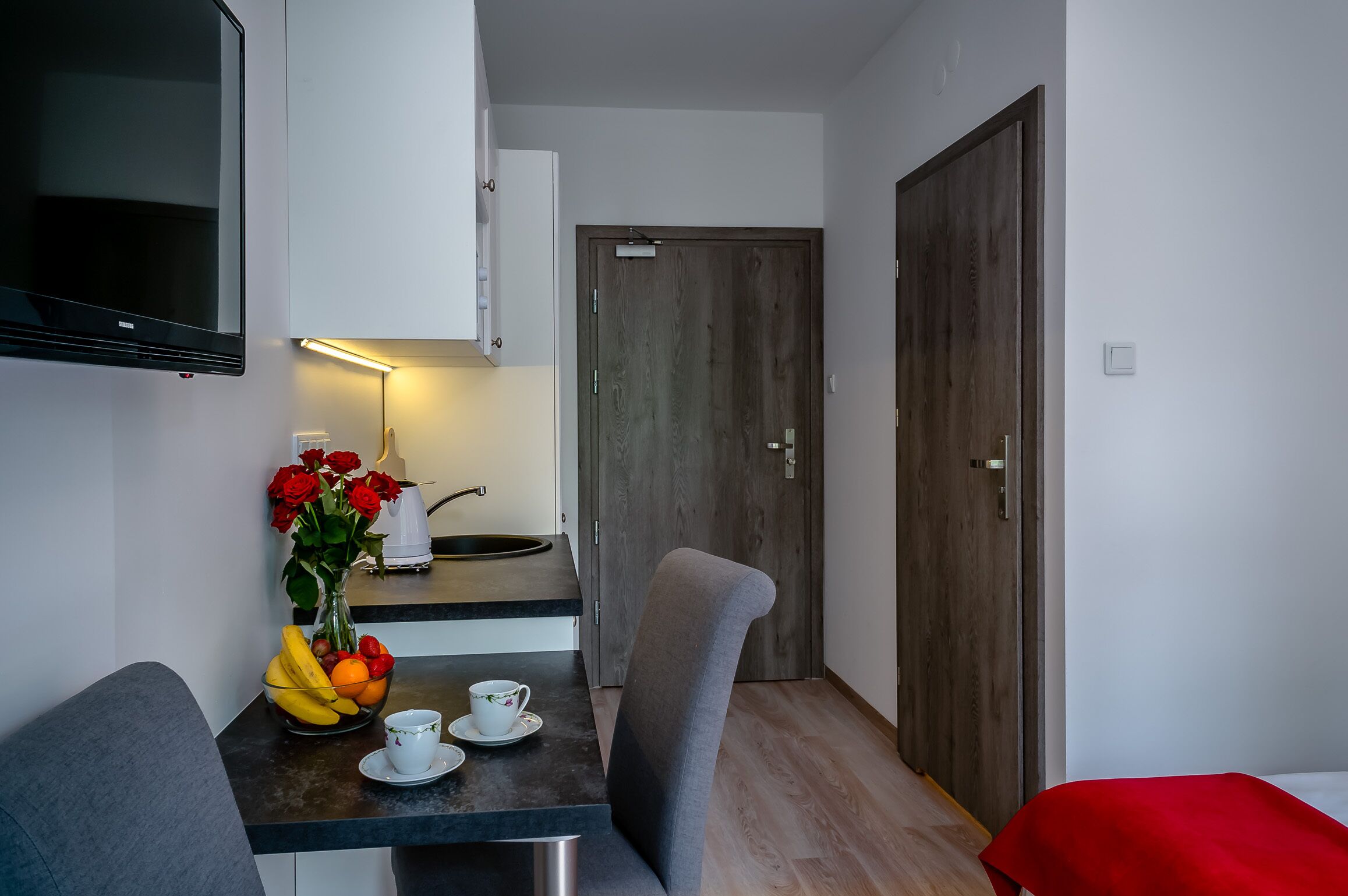 Een compacte kamer met kitchenette, een eethoek met tafel en stoelen, een aan de muur bevestigde televisie en een houten deur.