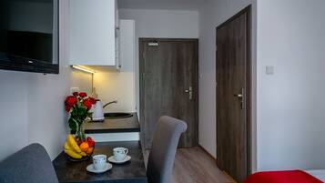 Een compacte kamer met kitchenette, een eethoek met tafel en stoelen, een aan de muur bevestigde televisie en een houten deur.