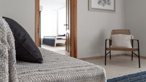 A room with a grey sofa, a wicker chair, a framed picture on the wall, and a doorway leading to another room.