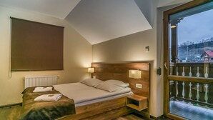 A bedroom with a wooden bed, a nightstand, a wall-mounted lamp, and a large window with a view of a building and trees.
