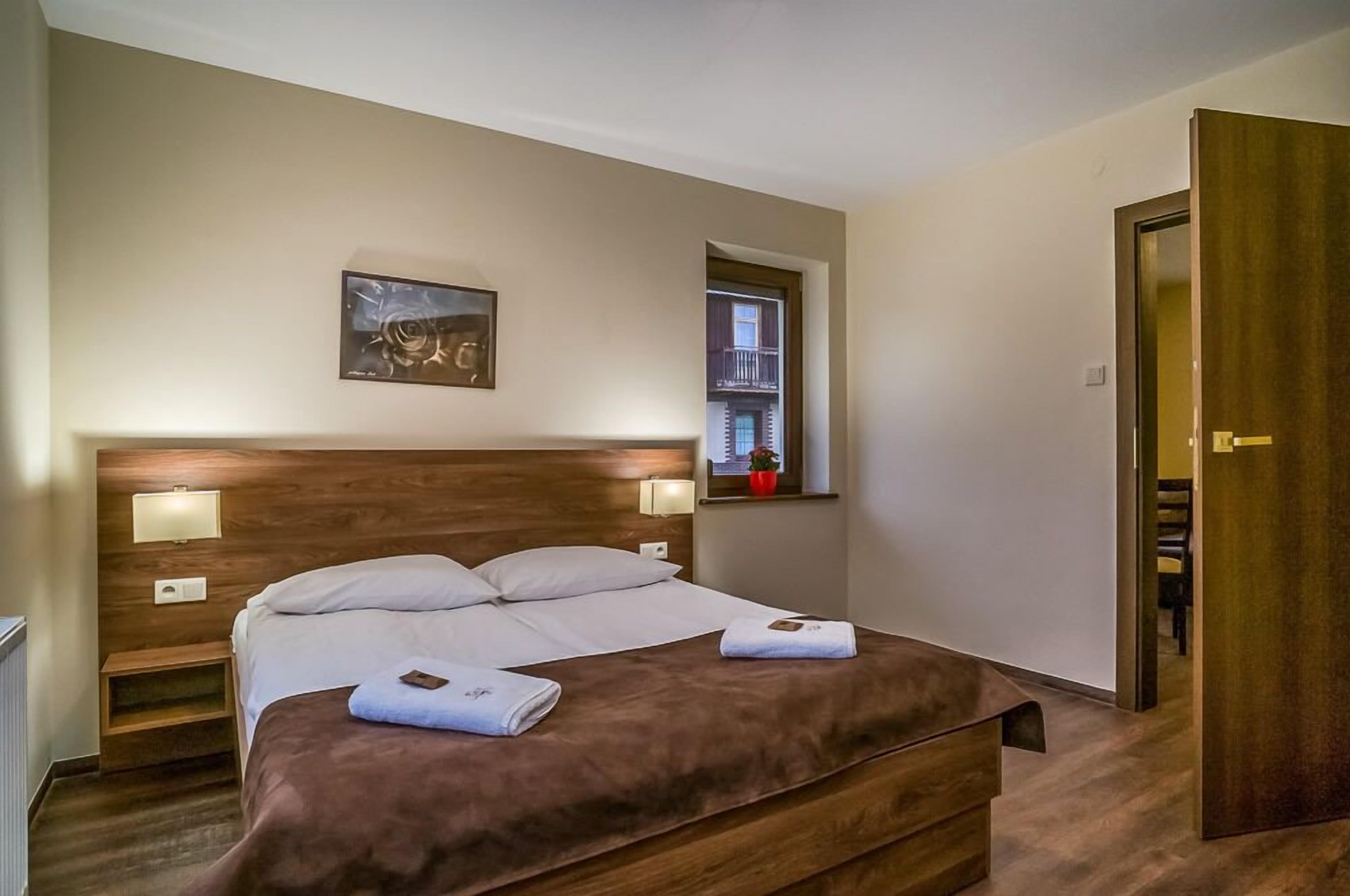 A hotel room with a large bed, bedside lamps, a small window with a view, and a door to the right.