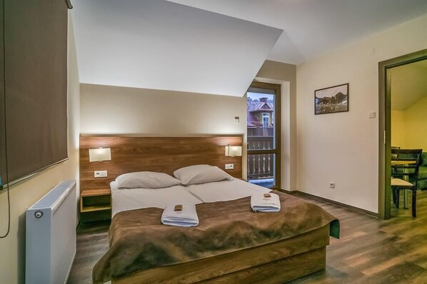 A bedroom with a wooden headboard, a bed with white linens, two bedside lamps, and a view of the outside through a window. - Apartamenty Eko (Szczawnica)