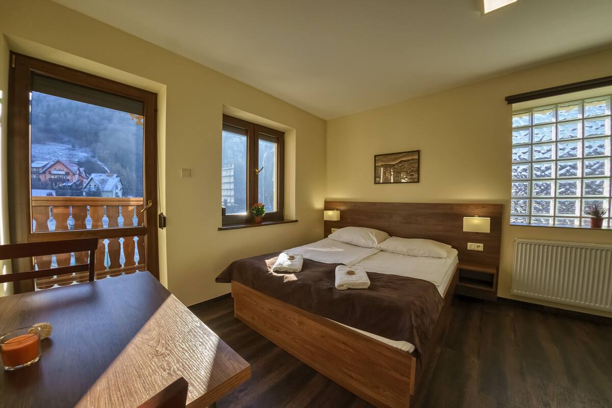 A bedroom with a wooden bed, a window with a view of houses, a small table with a candle, and a radiator.