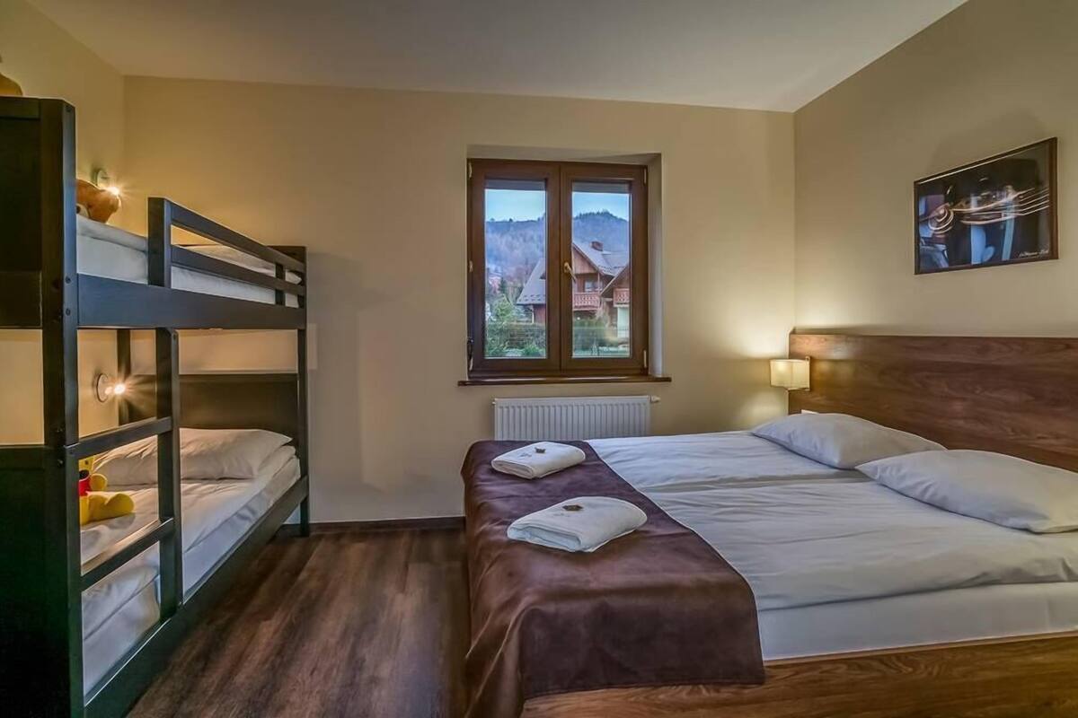 A room with a bunk bed, a single bed, a window with a view of houses and hills, and a painting on the wall.